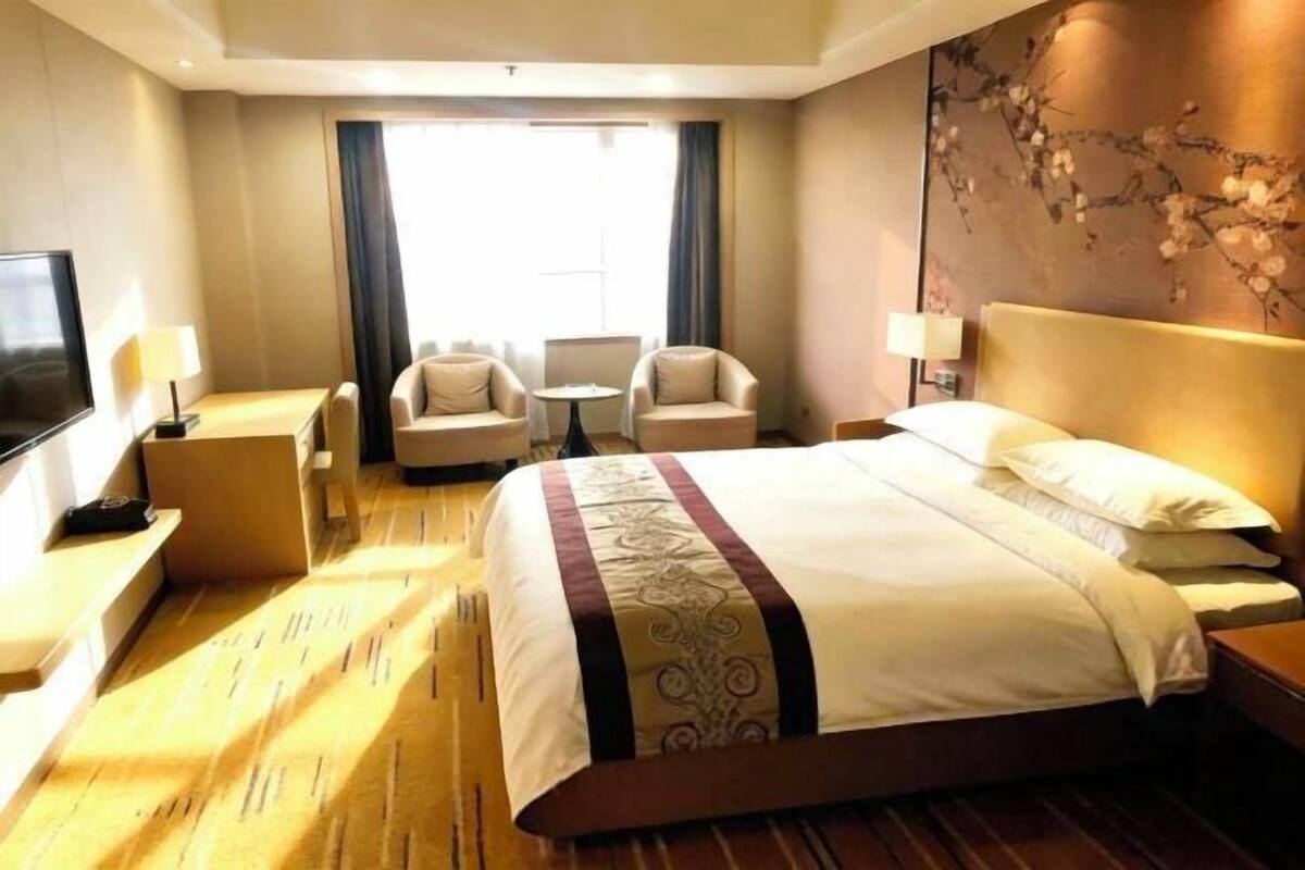 A hotel room with a large bed, two armchairs, a small table, a TV, and a window with curtains.