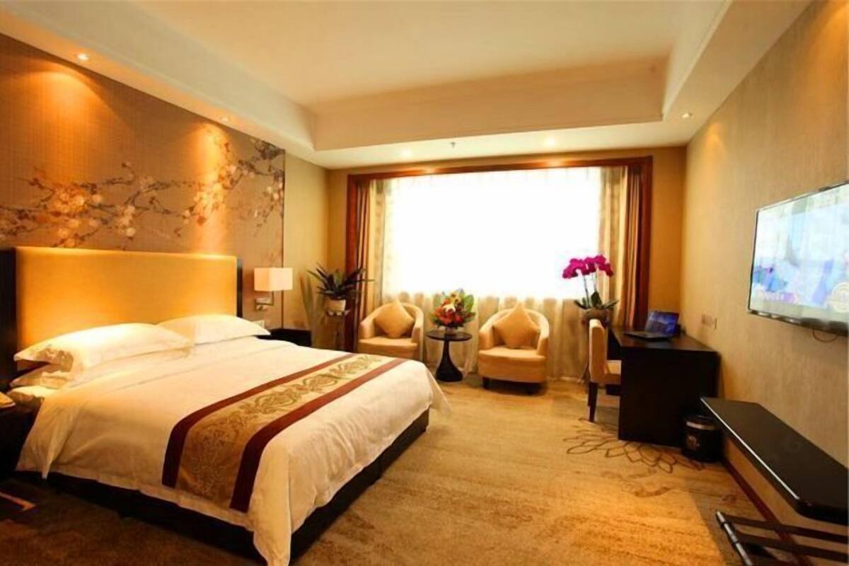 A hotel room with a large bed, two armchairs, a desk, a TV, and a window with curtains.