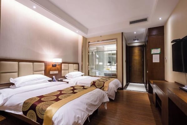 A hotel room with two beds, a television, a desk, and a bathroom visible through a glass door.