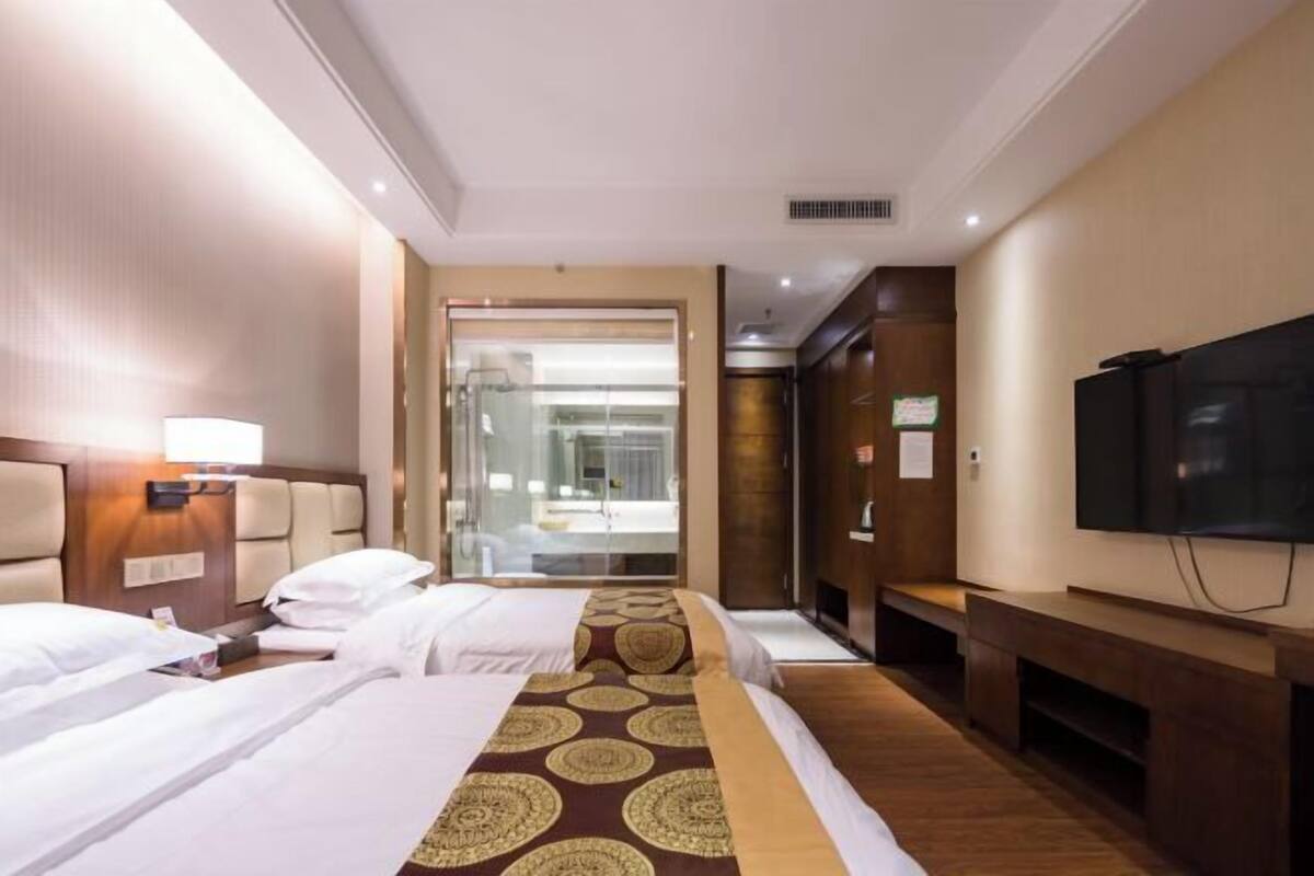A hotel room with two beds, a TV, a bathroom visible through a glass door, and a wooden floor.
