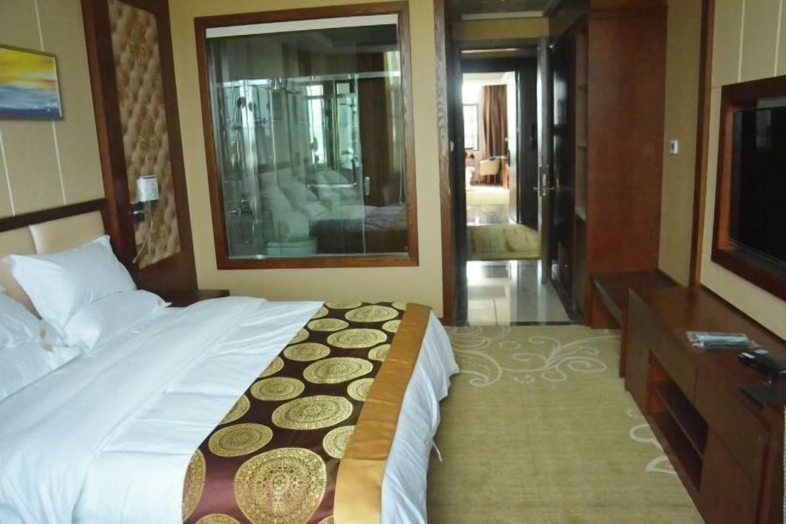 A hotel room with a large bed, a television, a bathroom with a glass door, and a view of the hallway.