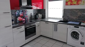 A compact kitchen with white cabinets, a red upper cabinet, a built-in oven, a washing machine, and a window with a view. - Vacation apartment with great views and hiking trails right outside the door (Burladingen)