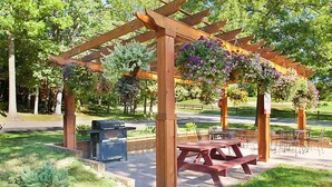 A wooden pergola with hanging flower baskets, picnic tables, and a barbecue grill in a park setting.