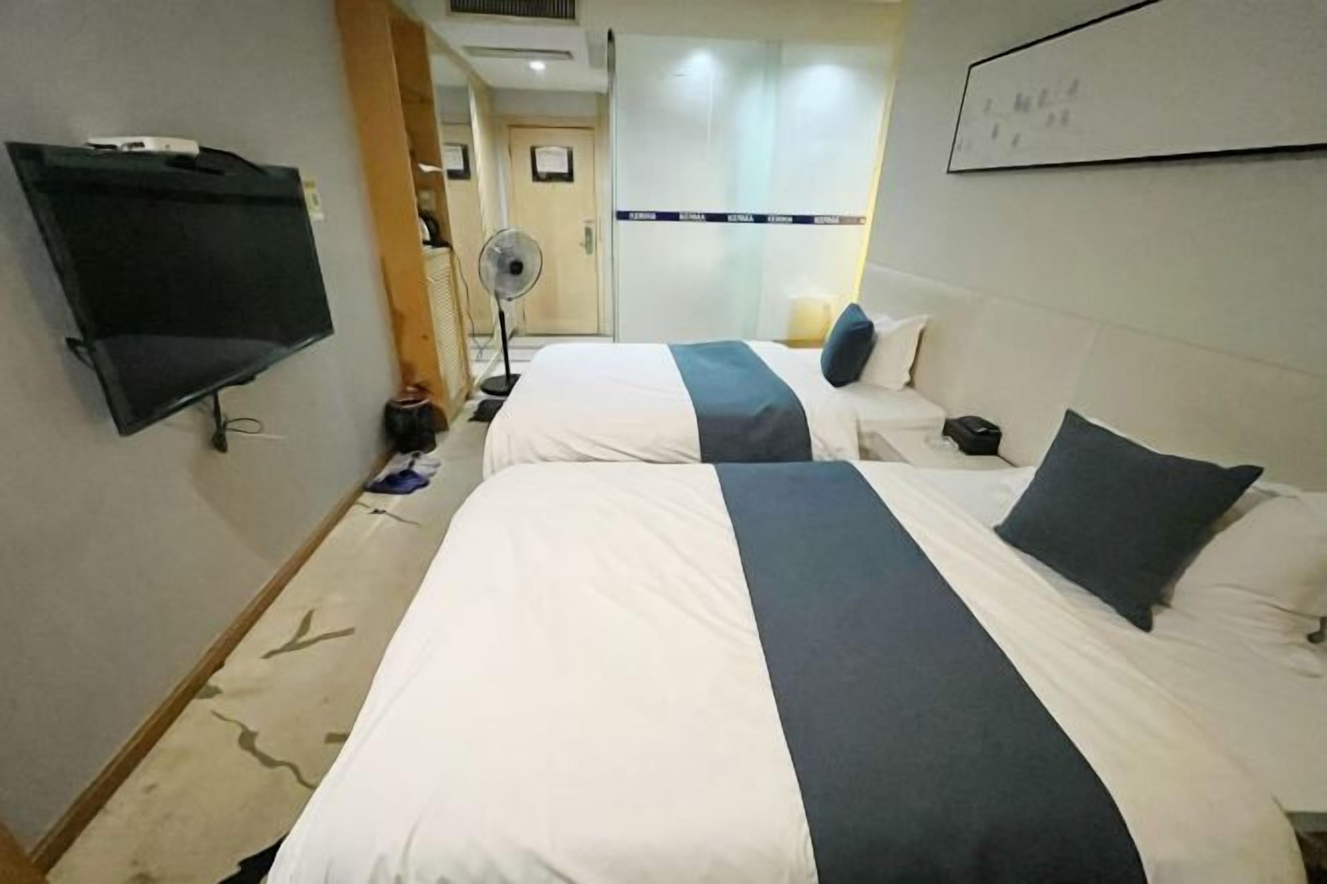 A hotel room with two beds, a flat-screen TV, a fan, and a whiteboard on the wall.