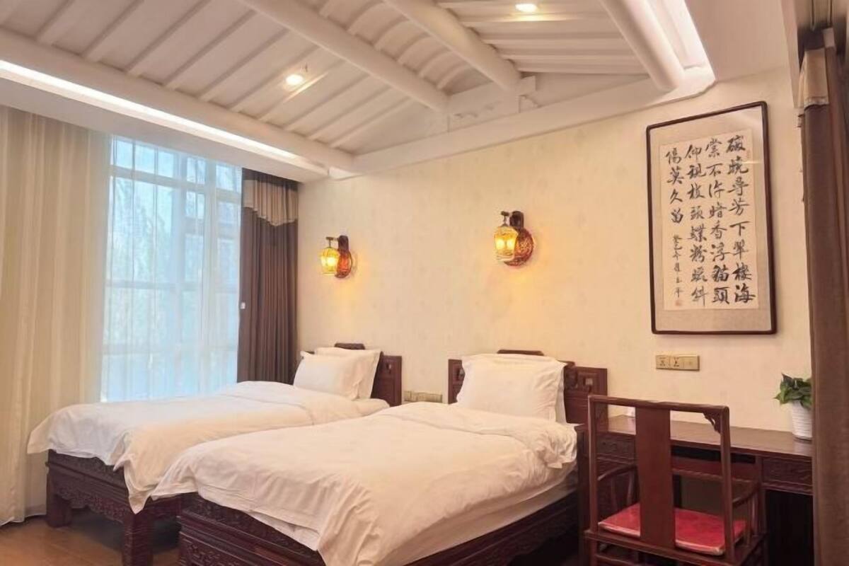 Une chambre d’hôtel avec deux lits, un bureau et une chaise. Un grand tableau calligraphié est accroché au mur.