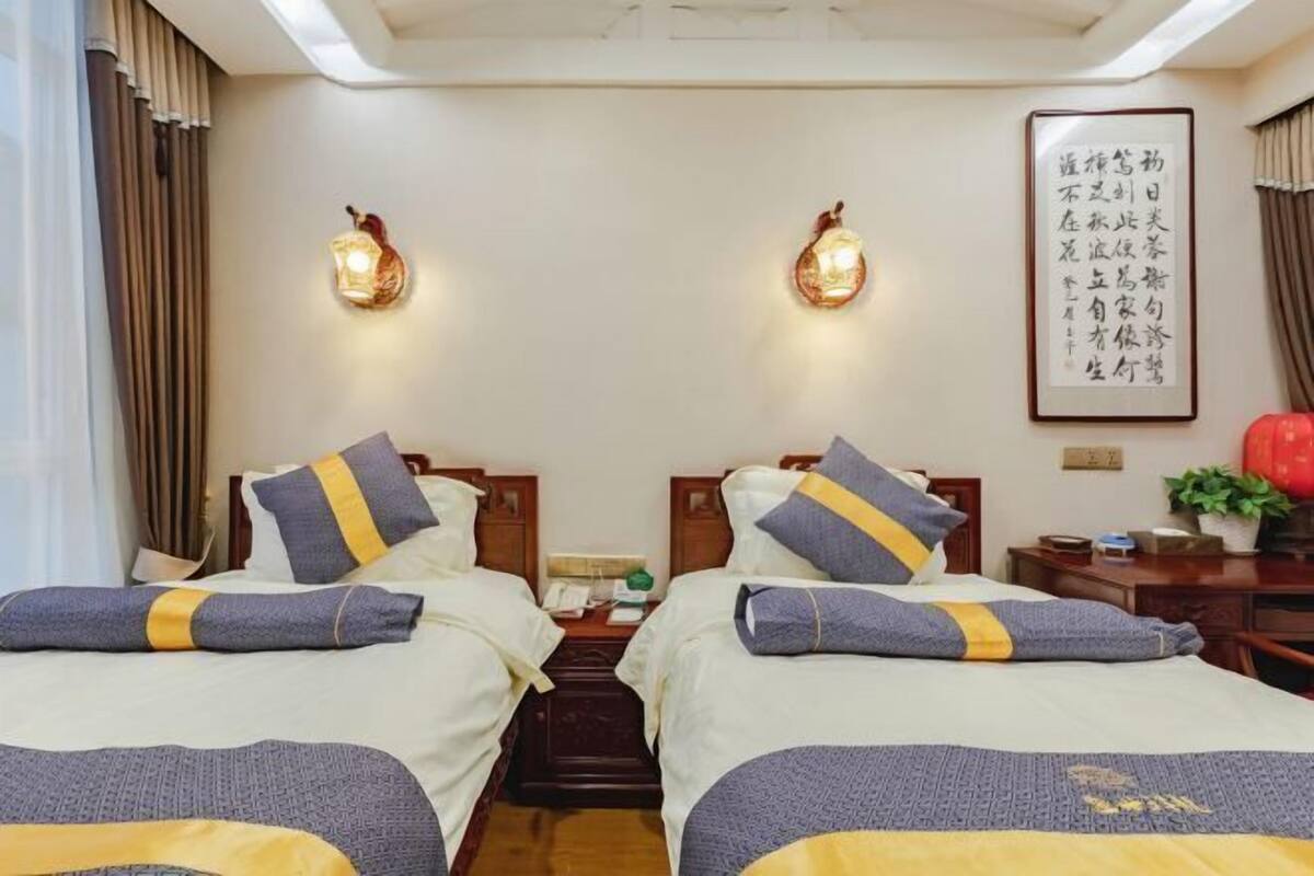 Une chambre d’hôtel avec deux lits, une table de chevet avec une lampe, une citation encadrée au mur et une plante en pot.