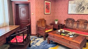 Ein traditionell eingerichtetes Zimmer mit Schreibtisch, Stühlen und einem roten Kissen. An der Wand hängt ein gerahmtes Kunstwerk und der Boden ist mit einem roten Teppich bedeckt.