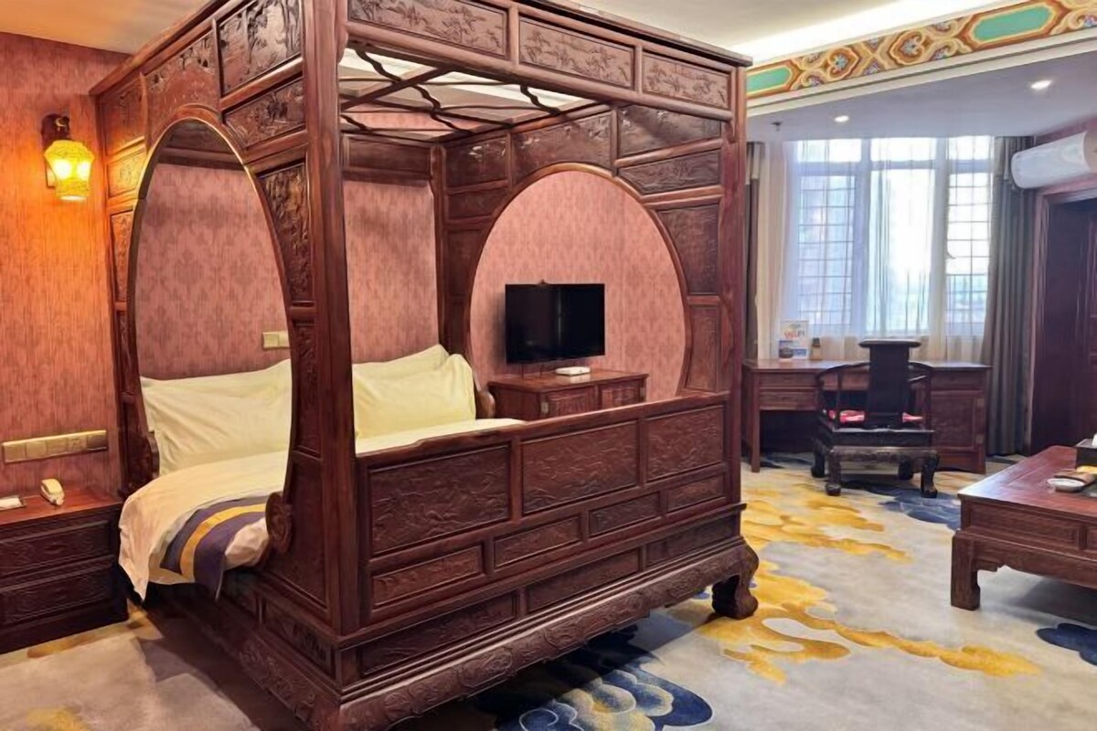 Uma cama de dossel tradicional com televisão em mesa de cabeceira de madeira, uma escrivaninha e uma cadeira em quarto com carpete estampado e janelas grandes.