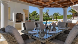 An outdoor dining area with wicker furniture, a glass table, and a view of a swimming pool and greenery. - Arundel - Javea, Costa Blanca (Javea)