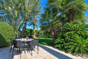 An outdoor patio with a round table and chairs, surrounded by lush greenery and palm trees.