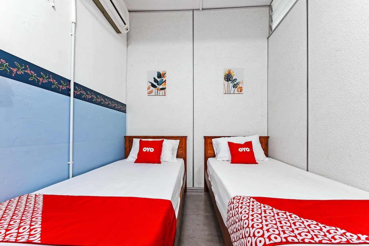 A room with two beds, each with a red and white patterned bedspread and a pillow with the word "OYO". The walls are painted blue and white, with floral artwork above the beds.