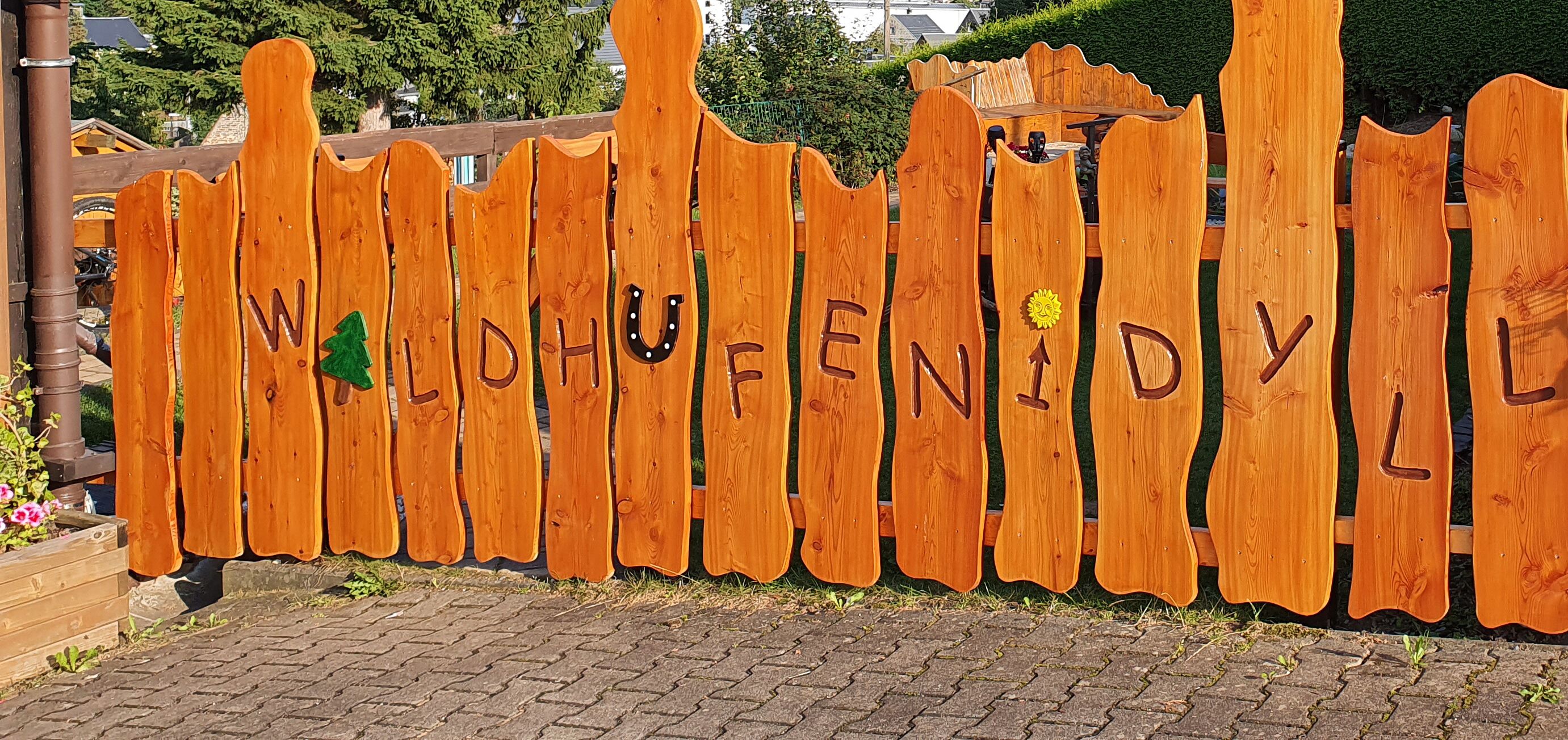 Ein Holzzaun mit eingekerbten Buchstaben, die ‚WALDHUFFENIDYL‘ buchstabieren.