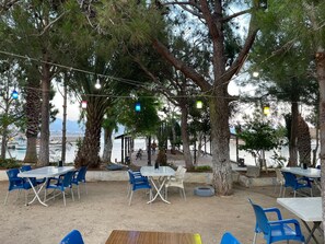 An outdoor seating area with tables and chairs, surrounded by trees and overlooking a body of water. - Ünlü Konaklama Kafe (Aydincik)