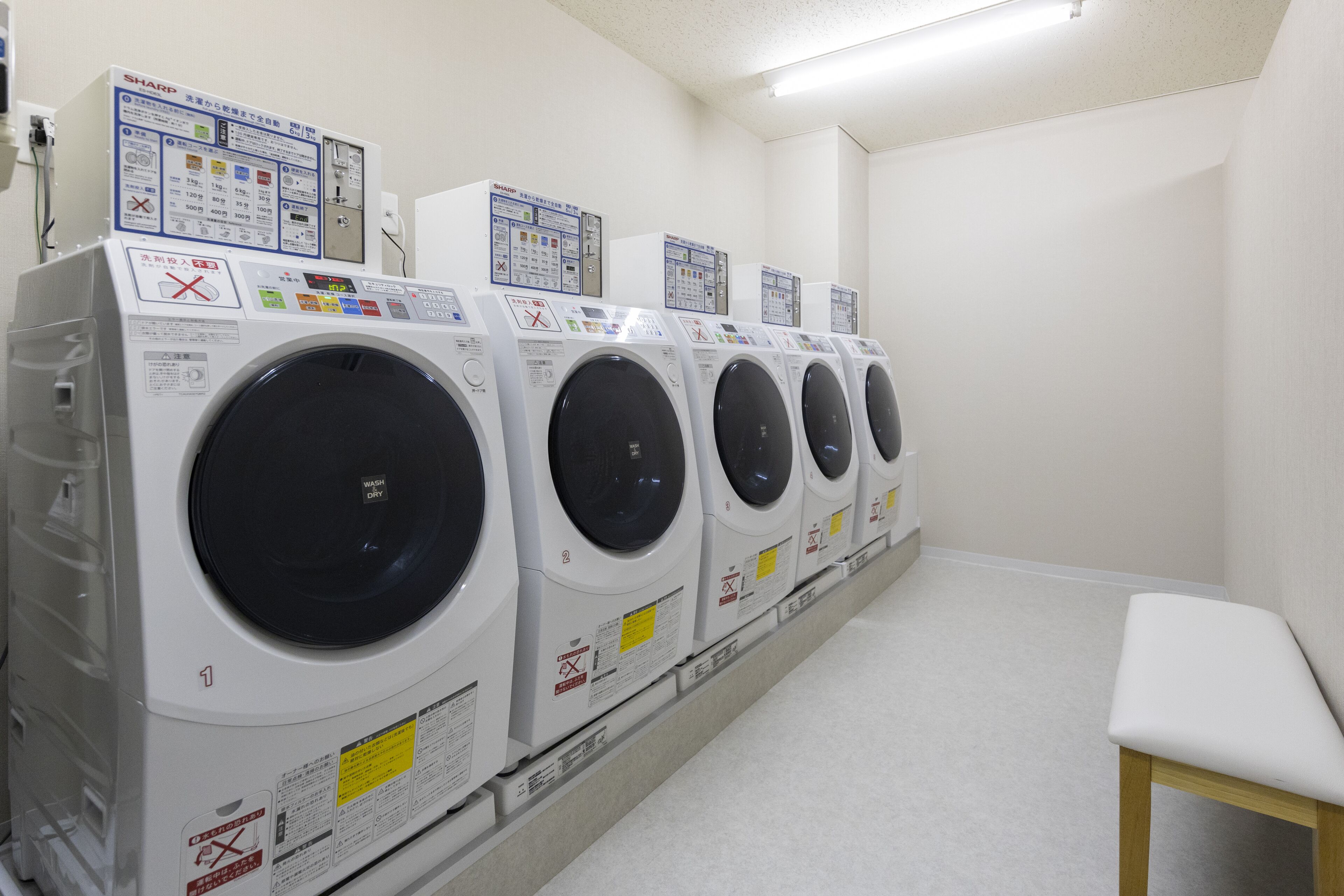 A row of white front-loading washing machines in a laundry room with a wooden bench.