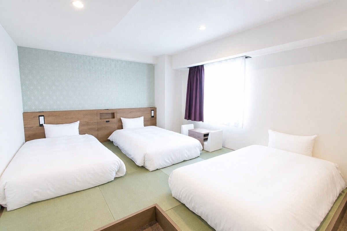 Habitación de hotel con tres camas, una mesita pequeña y un ventanal con cortinas.