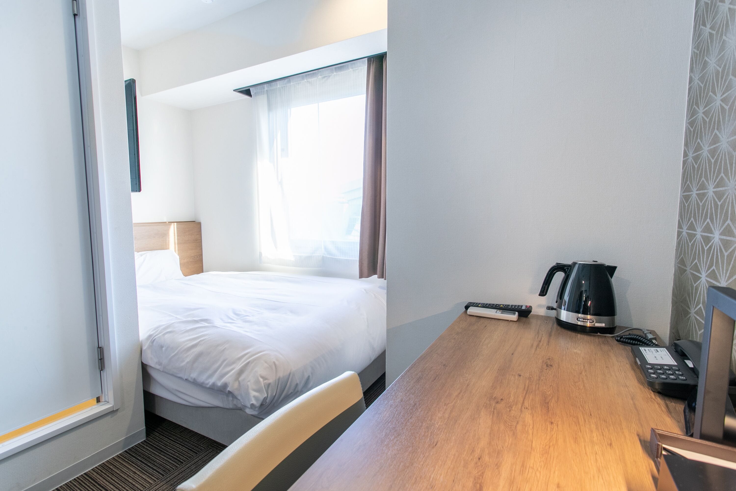 A hotel room with a bed, a wooden desk, a kettle, a telephone, and a window with curtains.