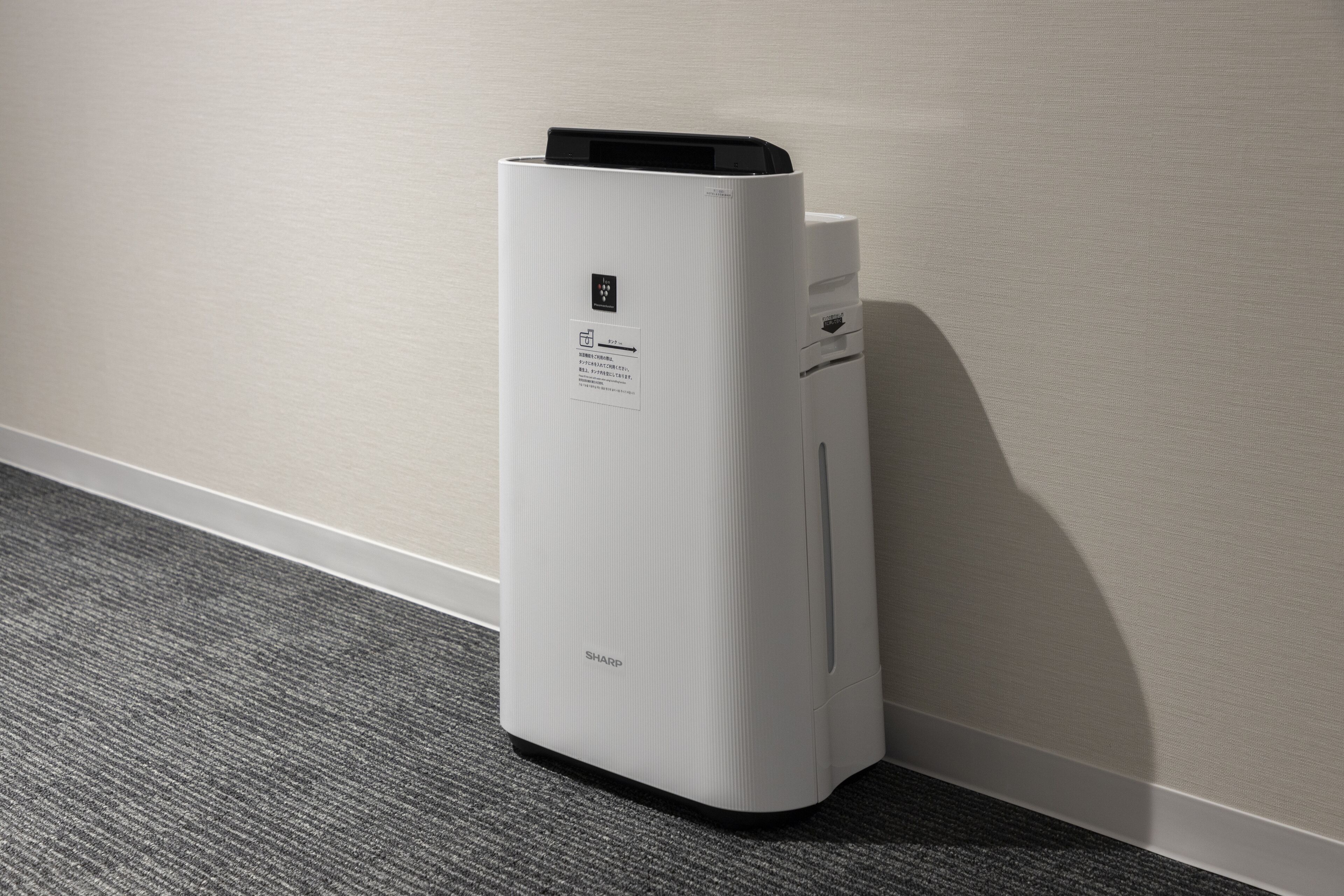 A white, modern, stationary air purifier with a control panel and a label on the side, placed against a wall on a carpeted floor.