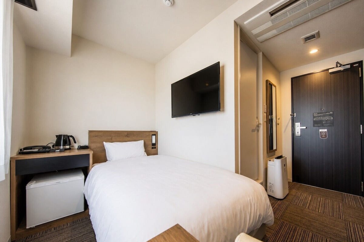 Habitación de hotel con cama, escritorio con cafetera, televisión y puerta con un cartel.