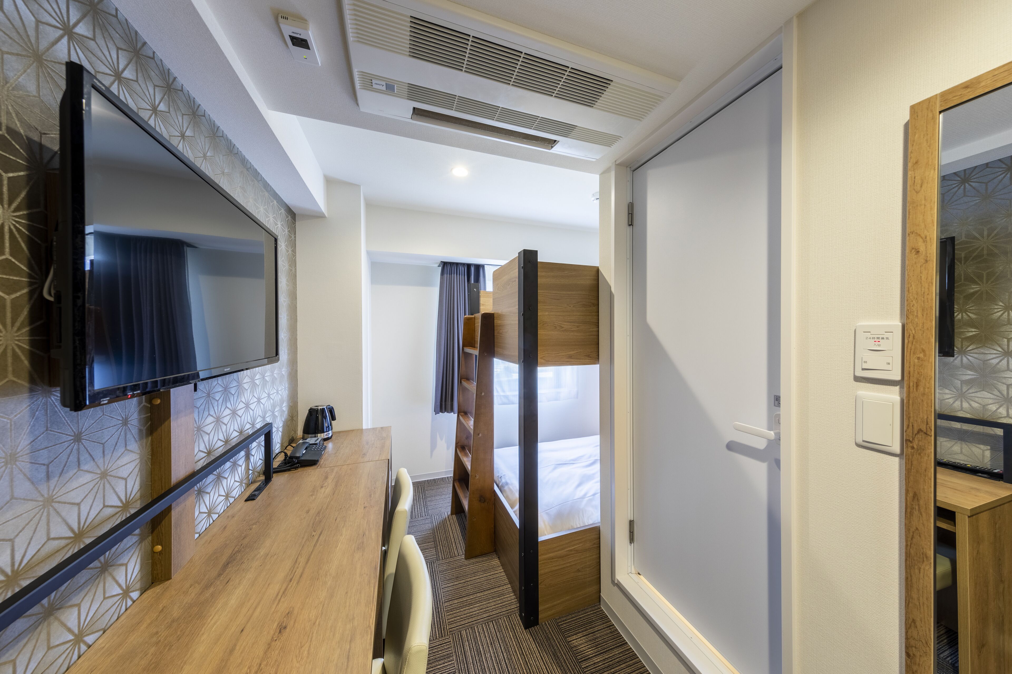 A compact hotel room with a bunk bed, a flat-screen TV mounted on the wall, a wooden desk with a chair, and a small table.