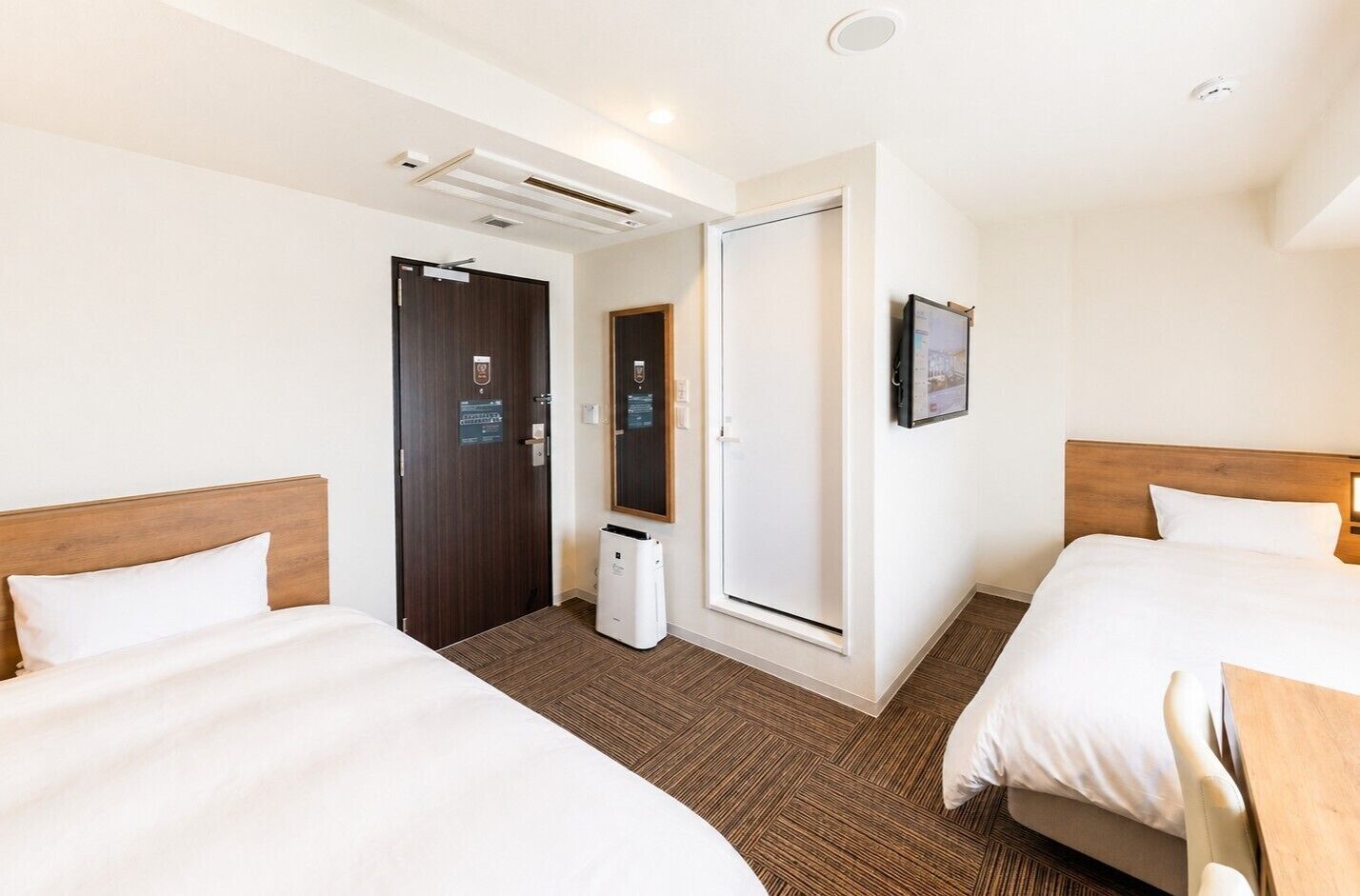 Une chambre d’hôtel avec deux lits, un petit bureau, un miroir et une porte donnant sur une salle de bain.