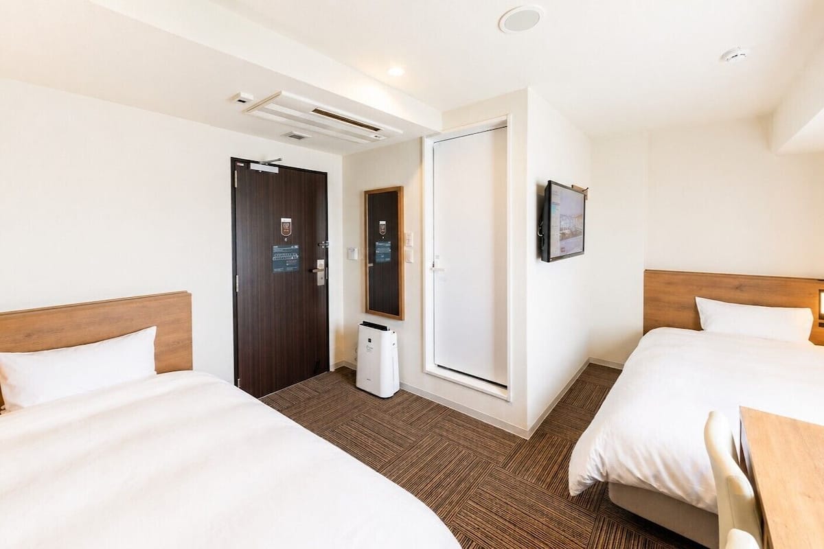 Hotellihuone, jossa on kaksi sänkyä, pieni työpöytä, peili ja ovi, joka johtaa kylpyhuoneeseen.