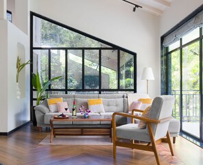 A living room with a sofa, a wooden chair, a coffee table with fruits and a vase, and a large window with a view of greenery. - Hearthstone By Getaway Stays (Alibag)