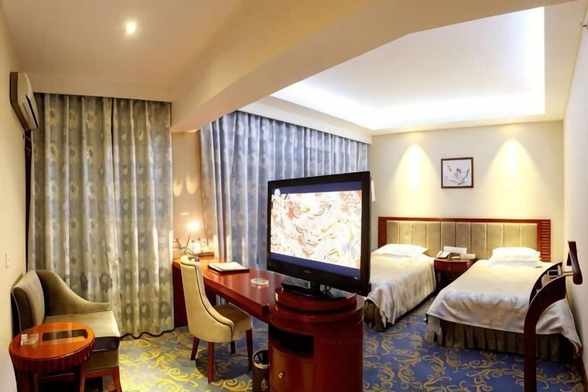 A hotel room with a TV, two beds, a desk with a chair, and a window with curtains.