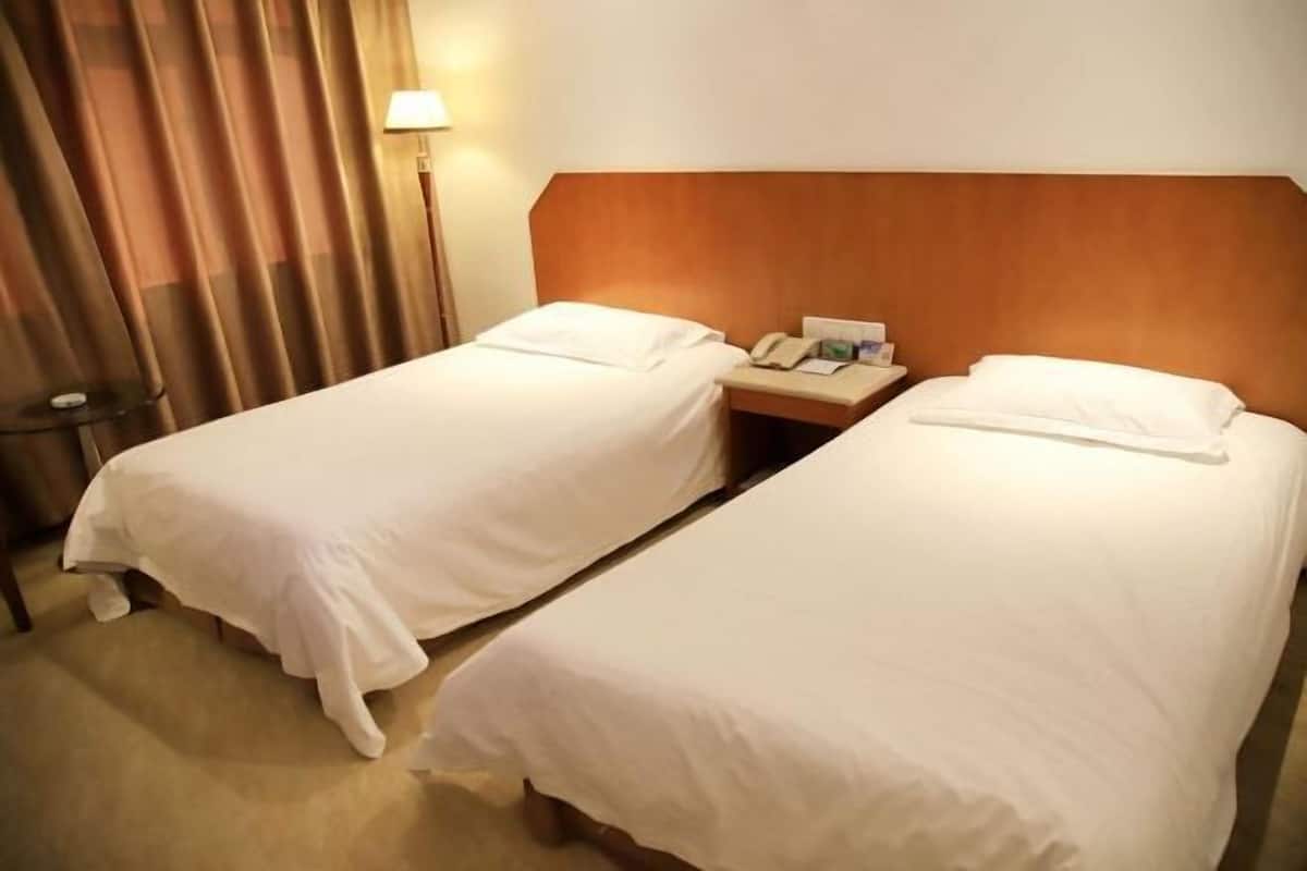 A hotel room with two single beds, a wooden headboard, a bedside table with a lamp, and a curtain.