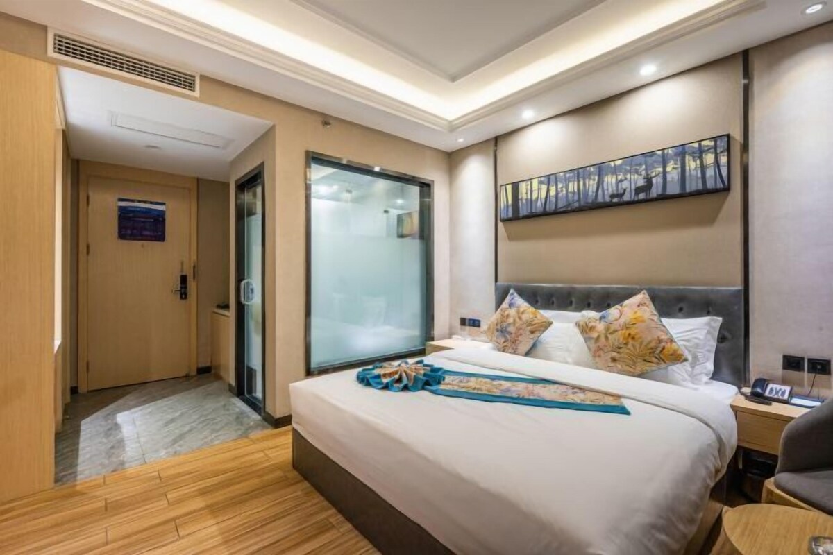 A hotel room with a large bed, a nightstand, a chair, a painting on the wall, and a door with a glass panel.