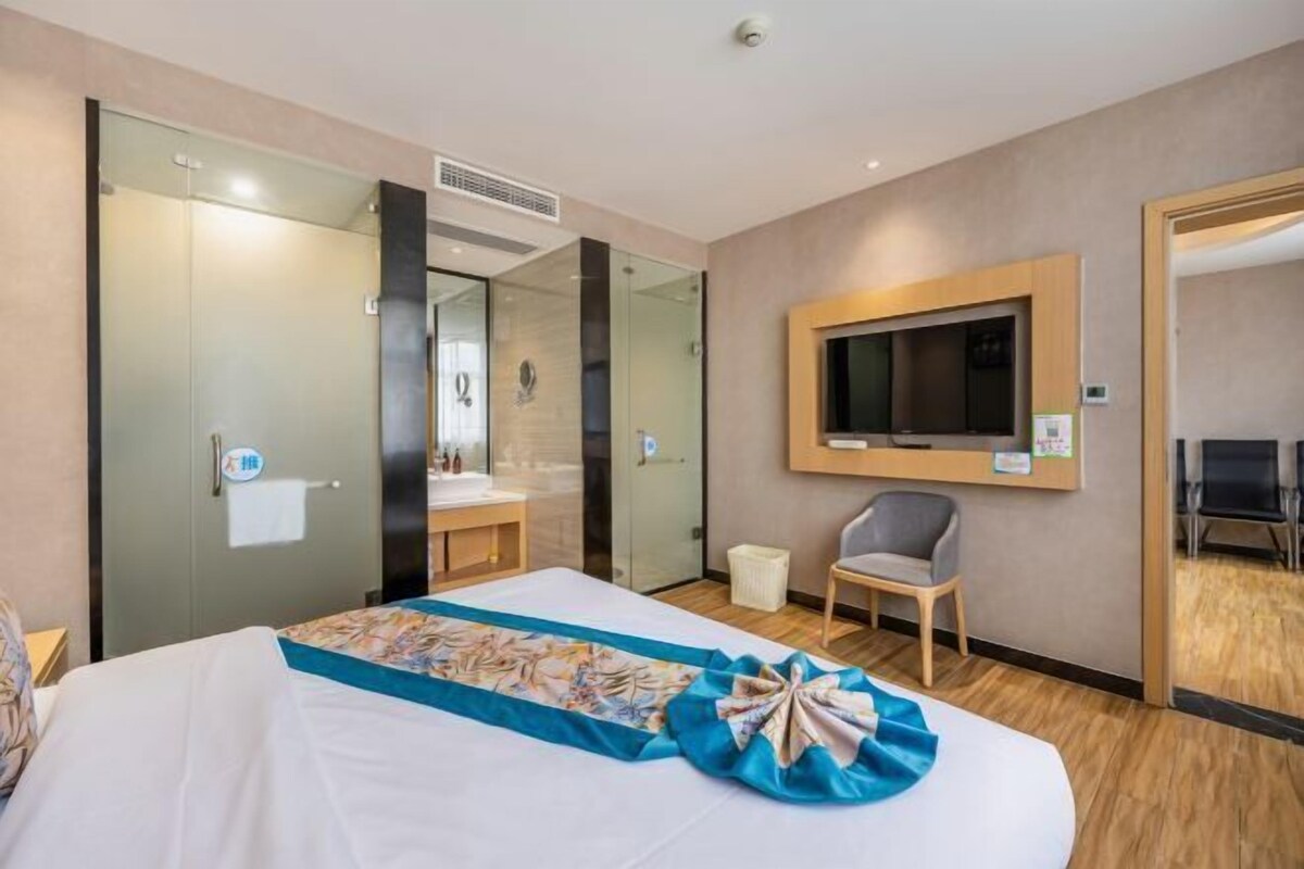 A hotel room with a bed, a desk, a chair, a TV, and a bathroom visible through a glass door.