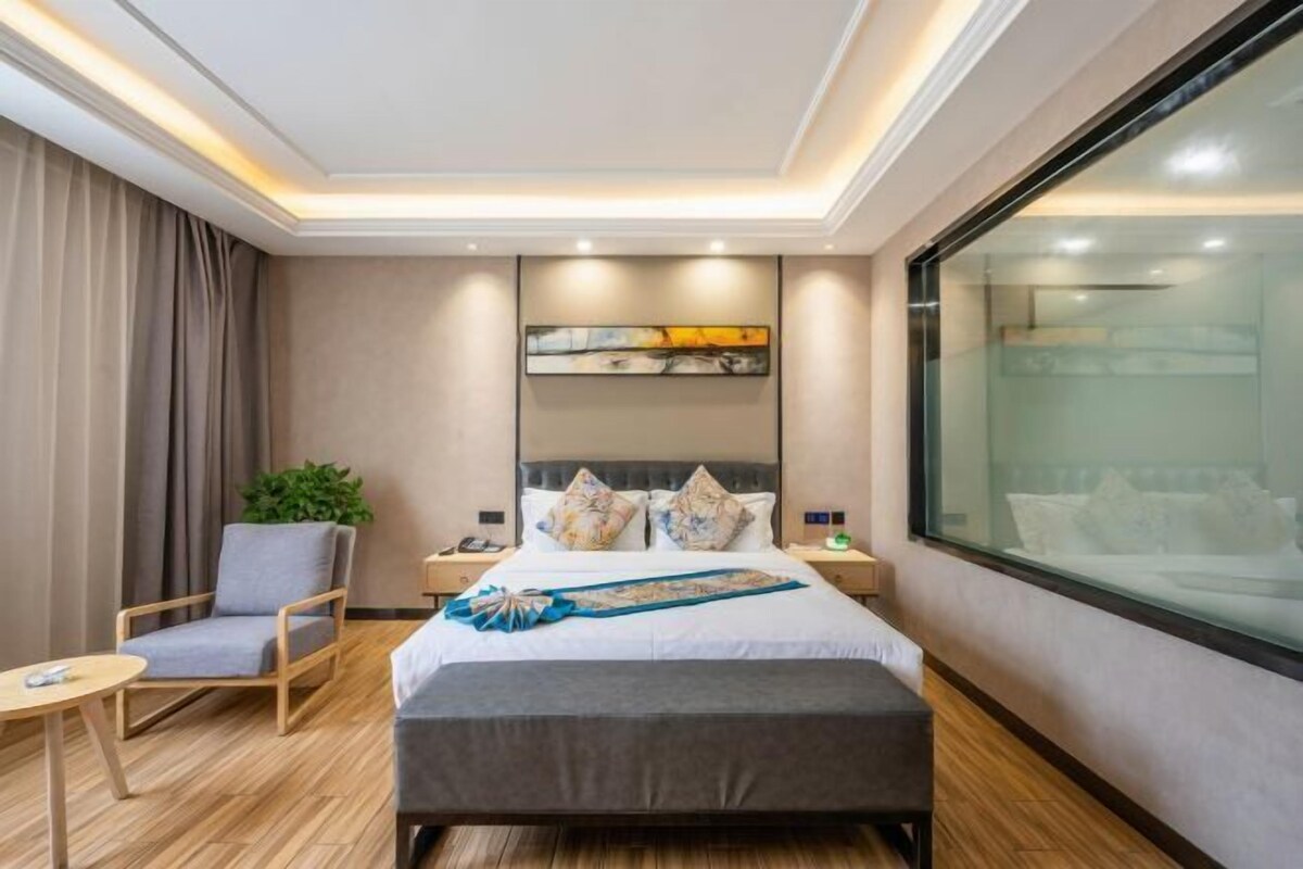 A modern hotel room with a bed, bedside tables, a chair, a plant, and a large window.