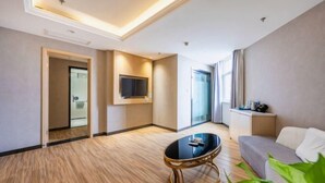 A modern hotel room with a wooden floor, a round glass coffee table, a grey sofa, a flat-screen TV mounted on the wall, and a large window providing natural light.