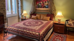 Un dormitorio con una cama grande, dos lámparas de noche, una mesita de noche, una silla, una ventana con cortinas y un cuadro en la pared.
