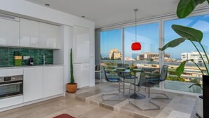 Uma cozinha moderna com armários brancos, uma mesa de jantar de vidro com cadeiras e uma grande janela com vista para a cidade.