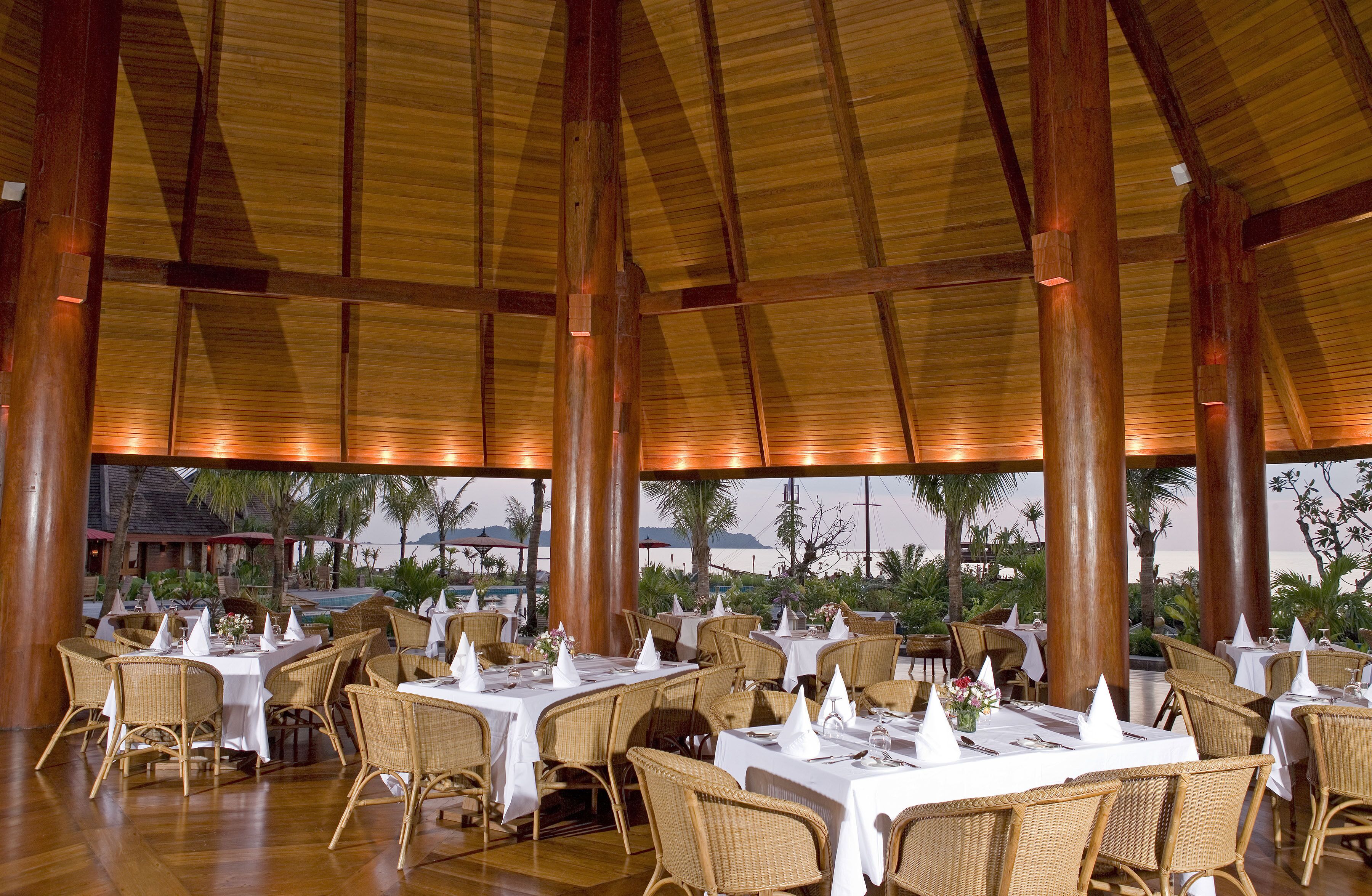 Un amplio comedor interior con techos altos y grandes vigas de madera, con mesas dispuestas con manteles blancos y sillas de mimbre, con vistas a un paisaje pintoresco.