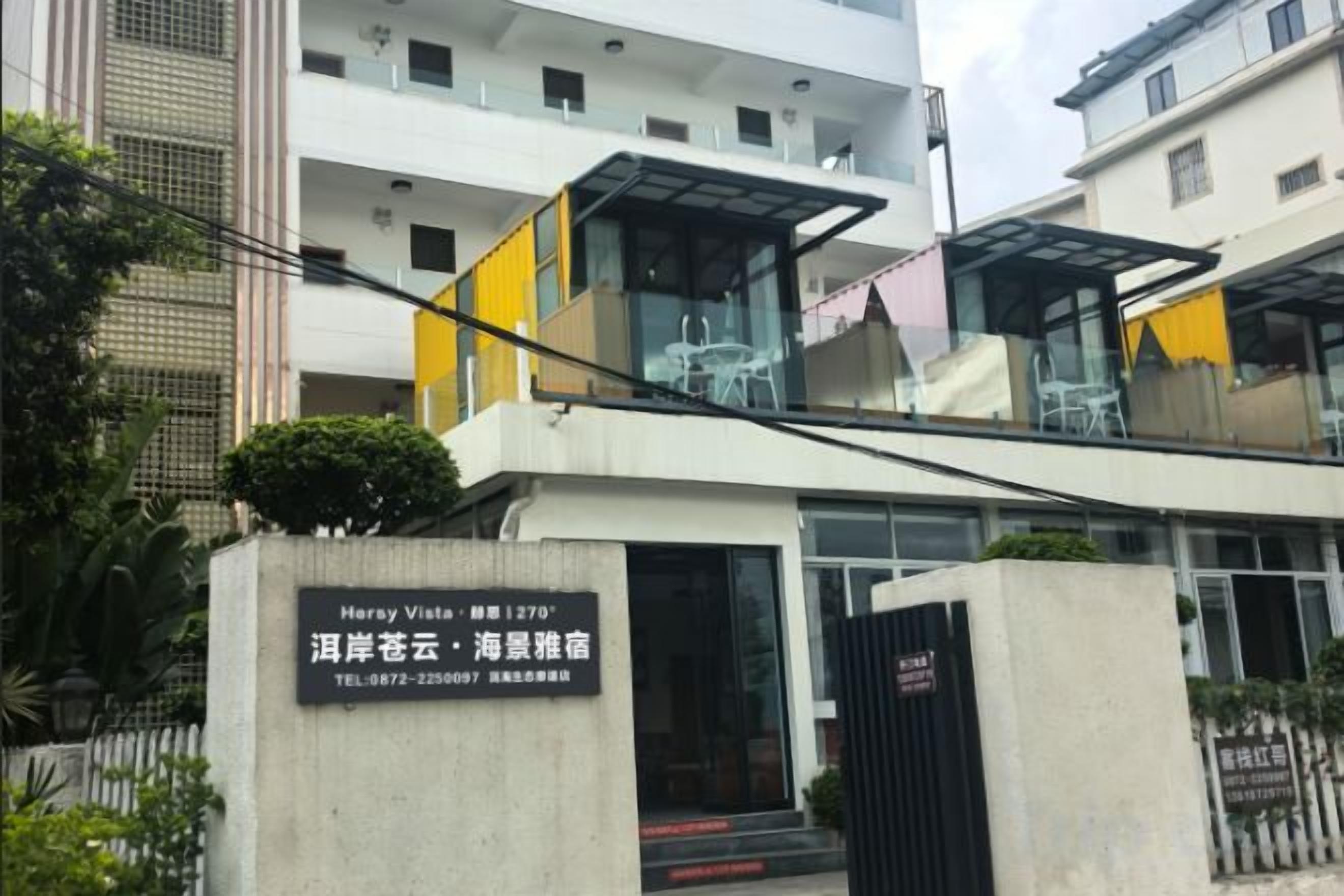 A modern hotel with a glass facade, a visible outdoor seating area, and a sign indicating the name and contact information.