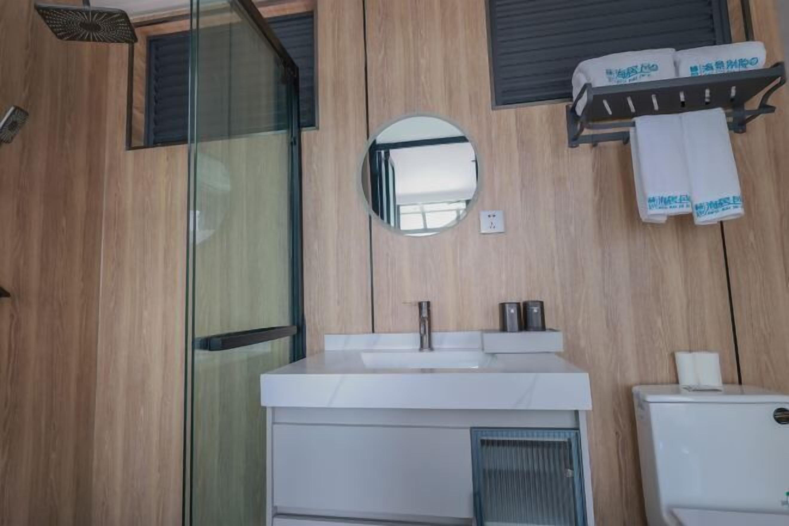 Un baño moderno con cabina de ducha de vidrio, un espejo redondo, un lavabo blanco y un perchero con toallas.