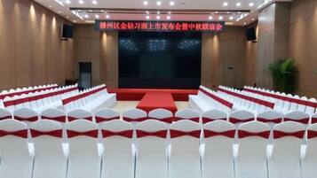 Un salón de conferencias con filas de sillas blancas, un escenario con alfombra roja y una pantalla grande.