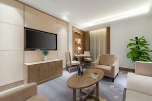 A modern hotel room with a flat-screen TV mounted on the wall, a wooden cabinet, a dining area with a table and chairs, and a large potted plant.