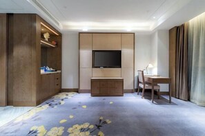 A hotel room with a flat-screen TV mounted on a wooden panel wall, a desk with a chair, a wardrobe, and a carpet with a floral pattern. - Chengtou Gangwan Hotel (Zunyi)