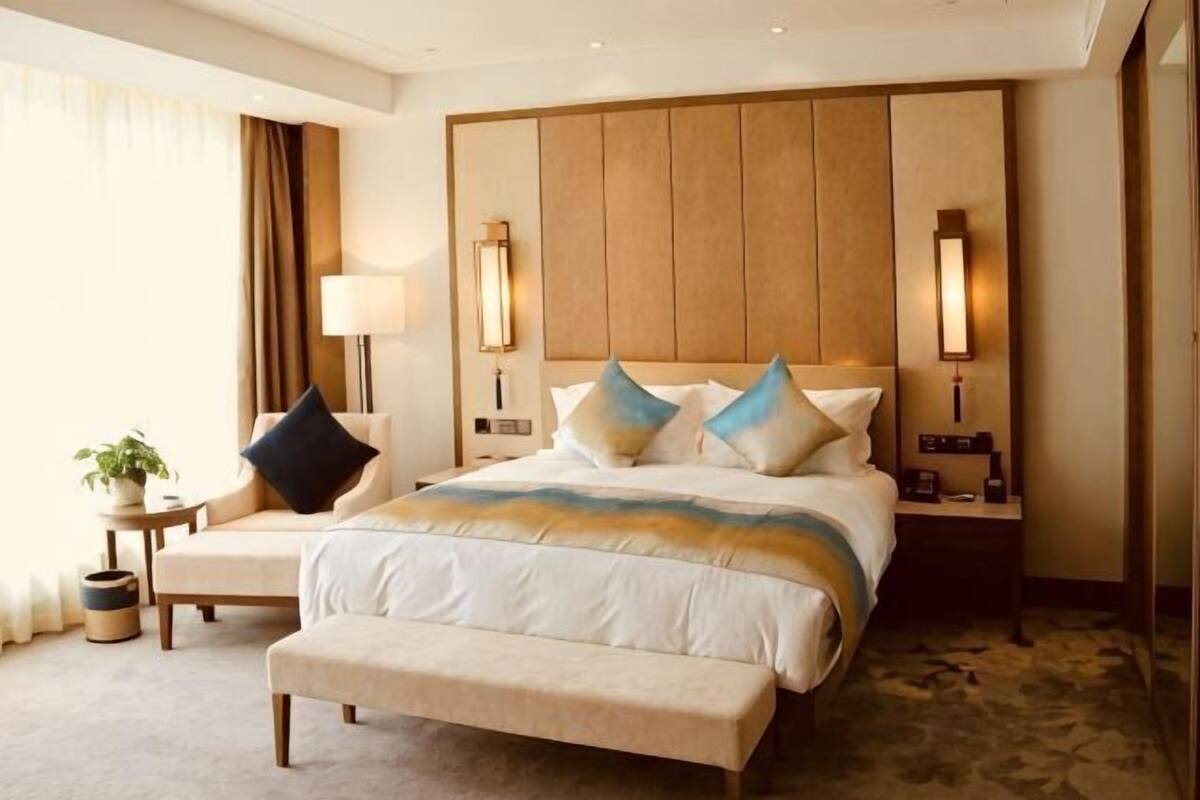 Ein Hotelzimmer mit einem großen Bett, Nachttischen, einer Bank, einer Lampe und einer Topfpflanze.