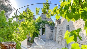 A stone building with a small door, surrounded by greenery and white chairs. - TD Trulli Locobello Stylish Cozy Trulli w Pool (Martina Franca)