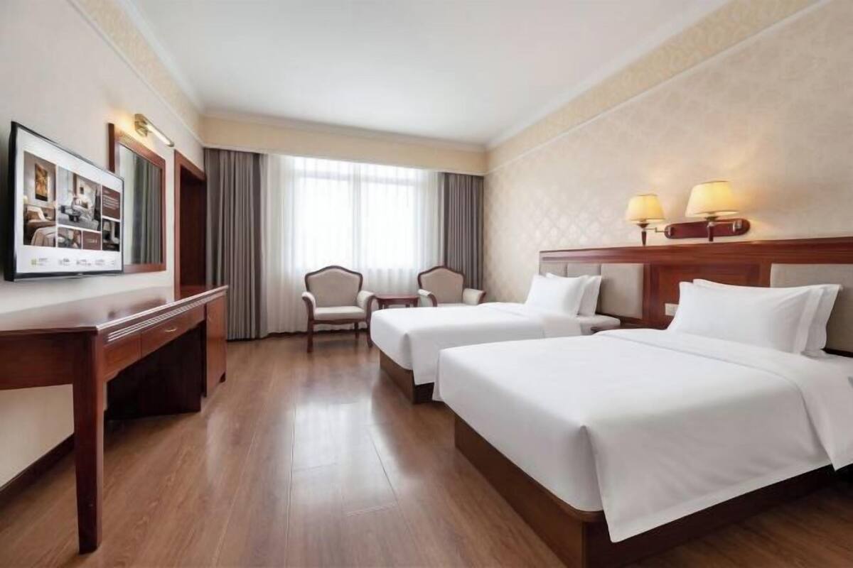 A hotel room with two beds, a desk, a chair, a TV, and a window with curtains.