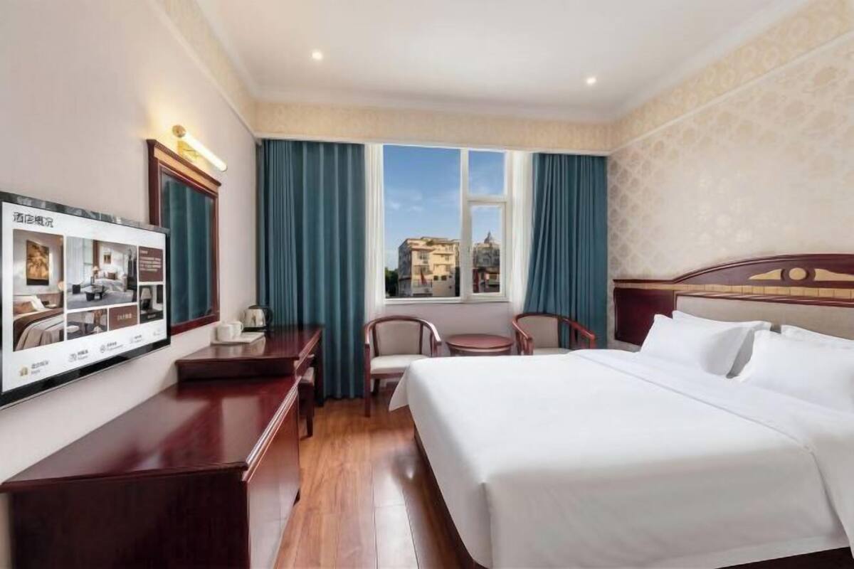 A hotel room with a bed, a desk, a chair, a small table, and a window with curtains.