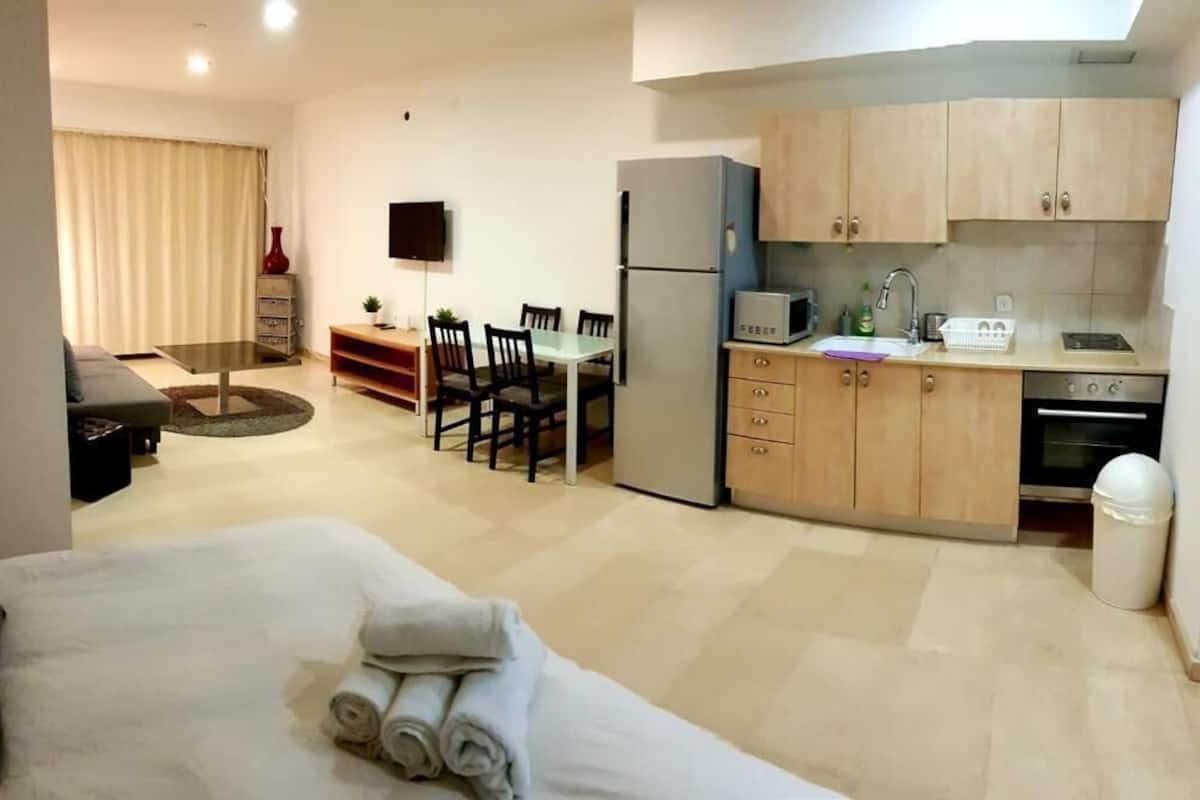 A compact hotel room with a bed, a small dining table, chairs, a kitchen area with cabinets and appliances, and a TV mounted on the wall.