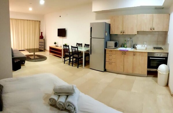 Una habitación de hotel compacta con cama, una pequeña mesa de comedor con sillas, kitchenette y televisor montado en la pared.