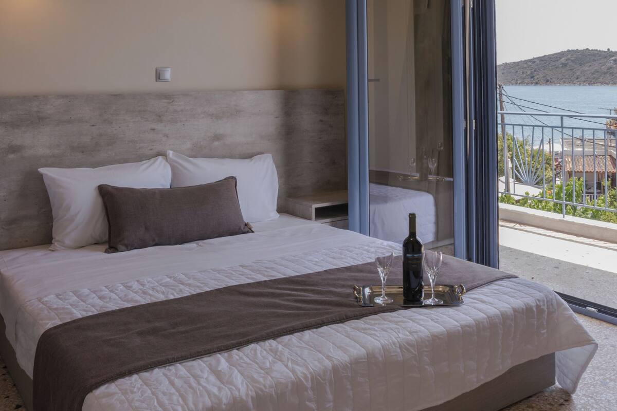 A bedroom with a large bed, a glass door leading to a balcony, and a view of the sea.