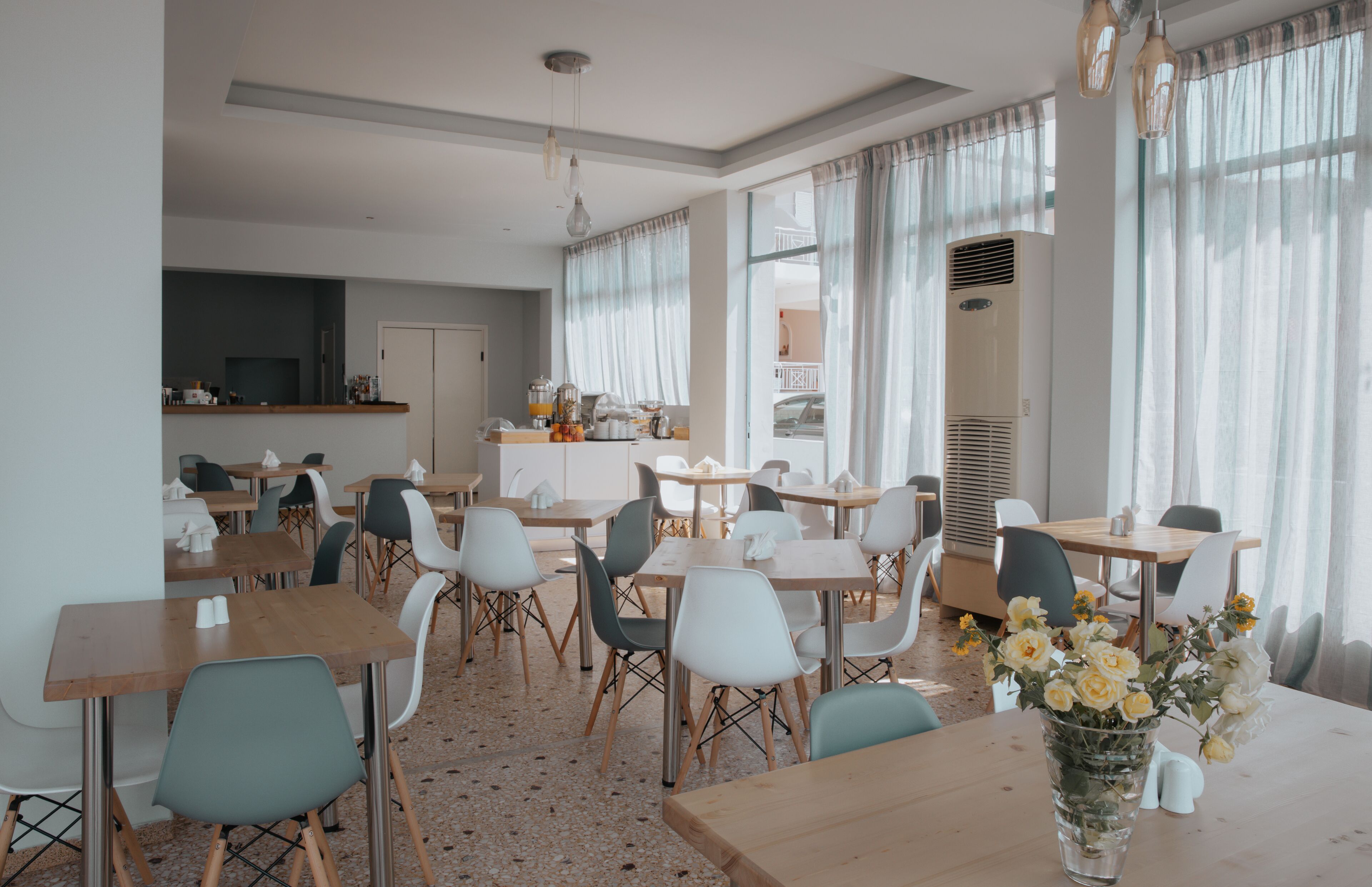 Uma sala de jantar com várias mesas e cadeiras, uma cozinha ao fundo e uma janela grande com cortinas.