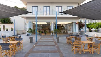 Un patio al aire libre con mesas y sillas de madera, plantas en macetas y un edificio con varios pisos y balcones.