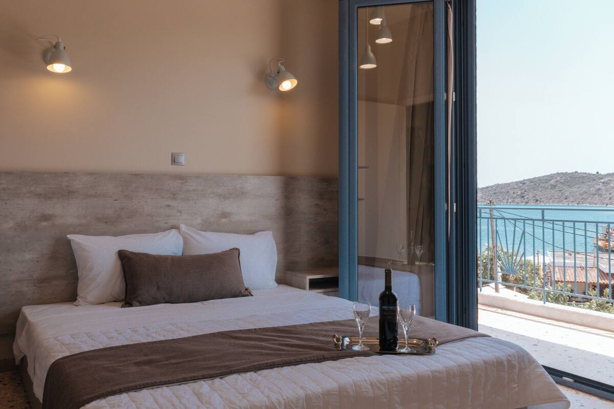A bedroom with a bed, pillows, a bedside table with a bottle and glasses, and a view of the sea through a balcony.